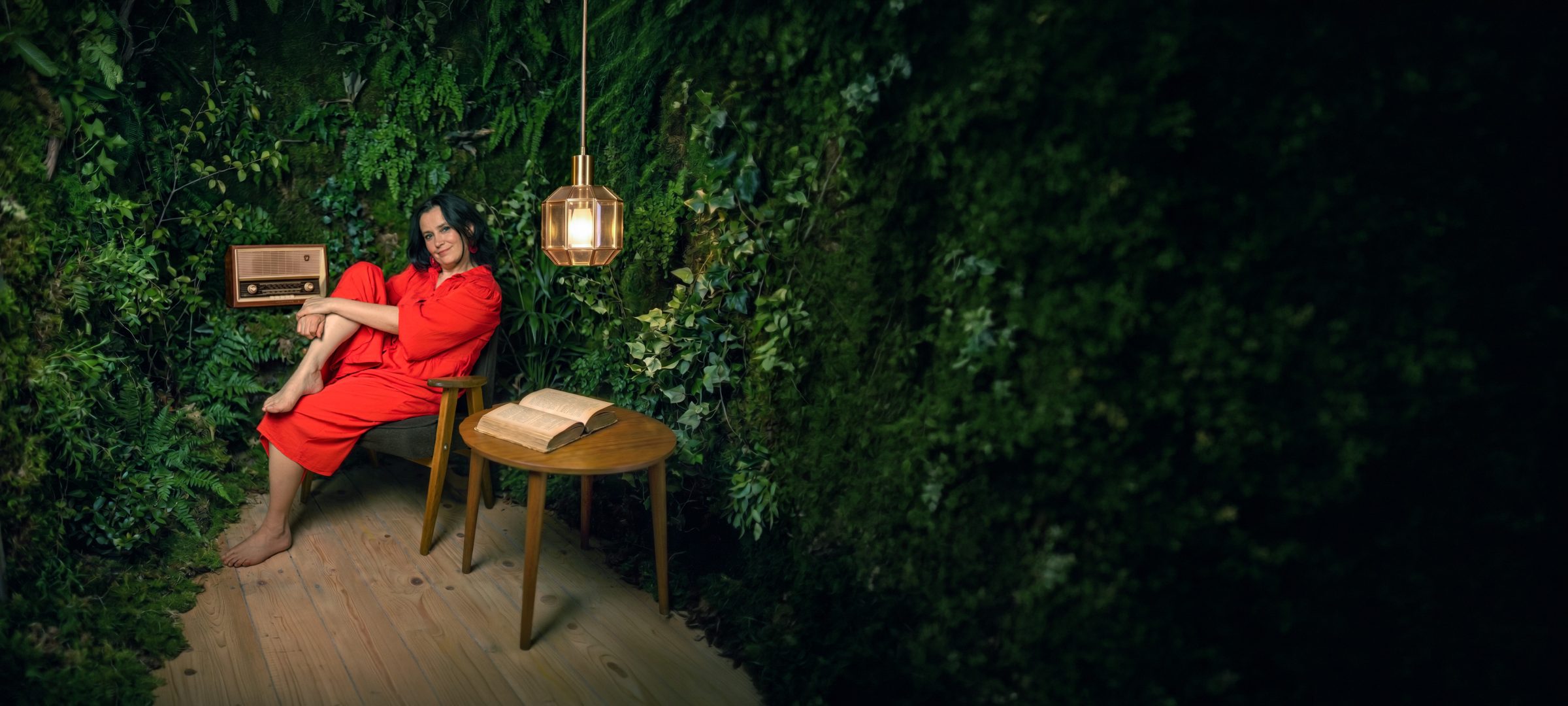 Karolina Tuz w czerwonej sukience, w fotelu wśród zieleni, obok lampa i stary radioodbiornik
